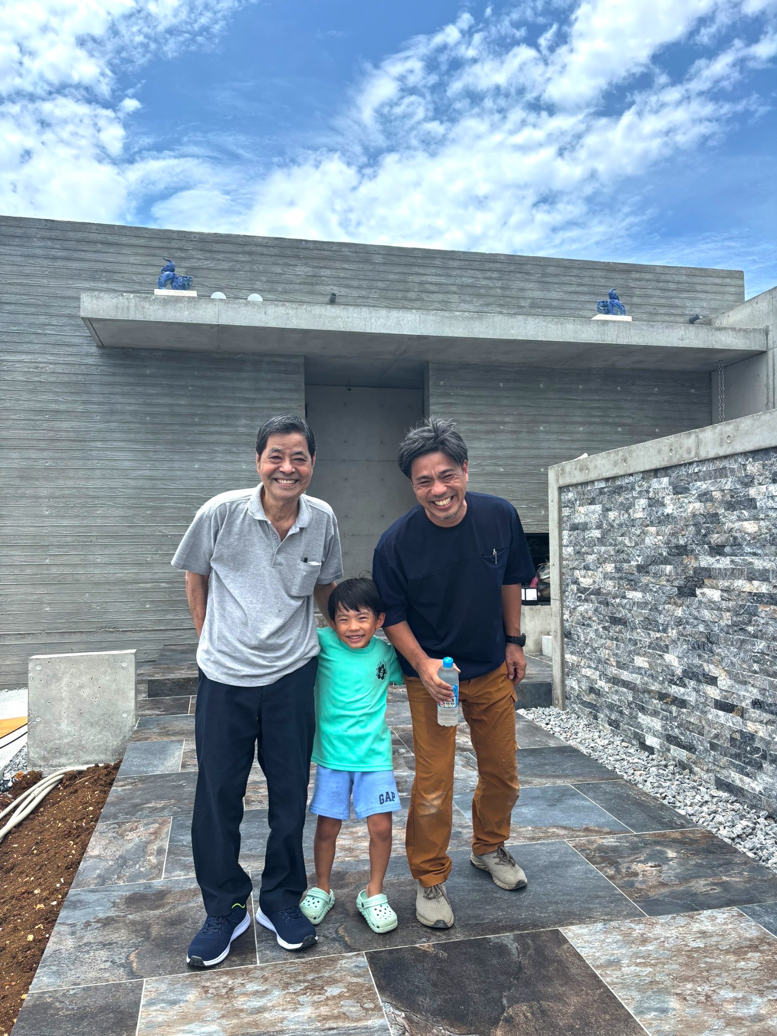 父が制作した青いシーサーを設置した建物の前に並ぶ、父・新垣信一、息子・新垣信人、孫の三代